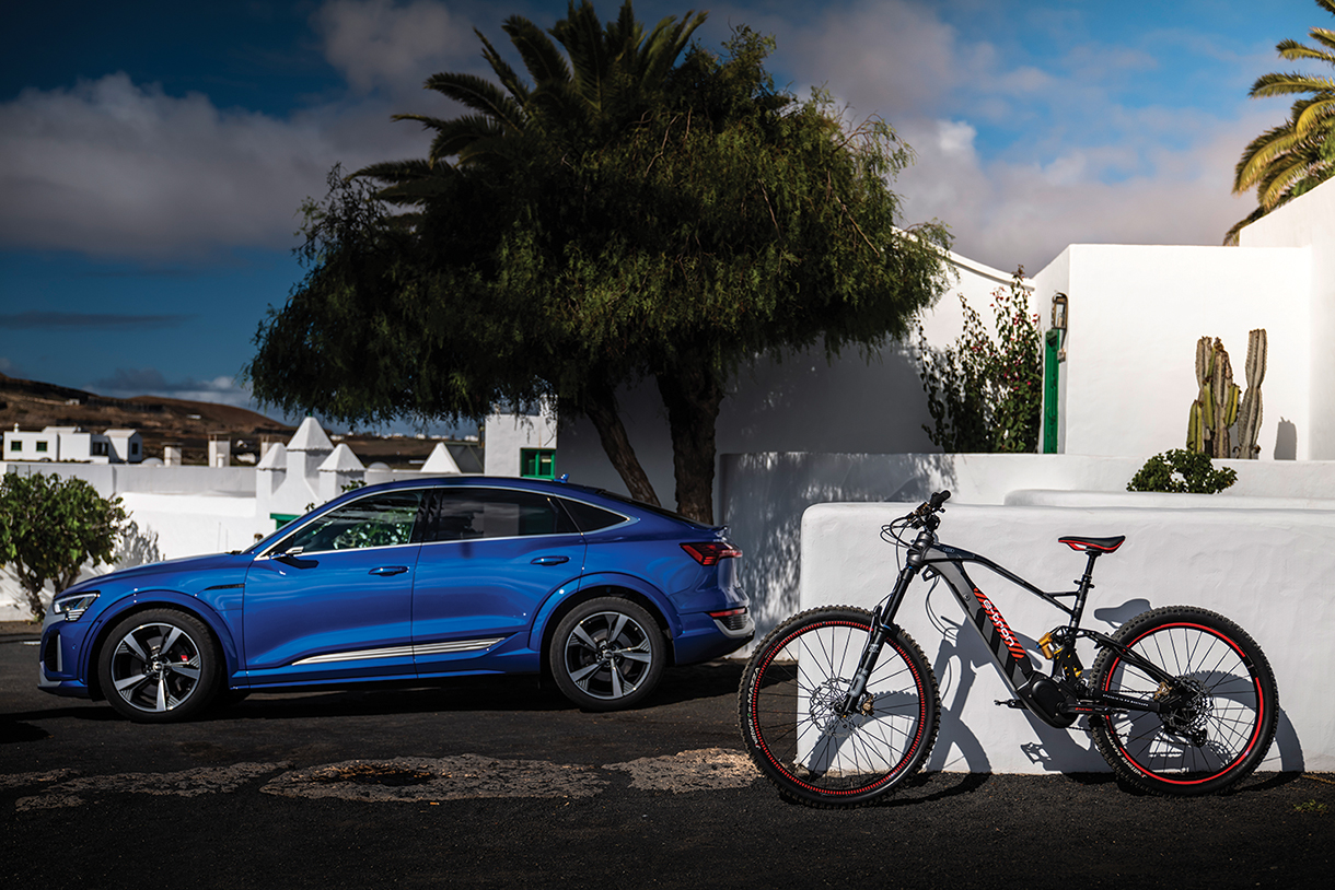 The Audi eMTB electric mountain bike in gray and red takes inspiration from its Rally Paris-Dakar racing car. Its e-tron branding is prominently showcased on the battery. The bike is parked in front of white building with a blue Audi SQ8 Sportback e-tron electric SUV next to it. 